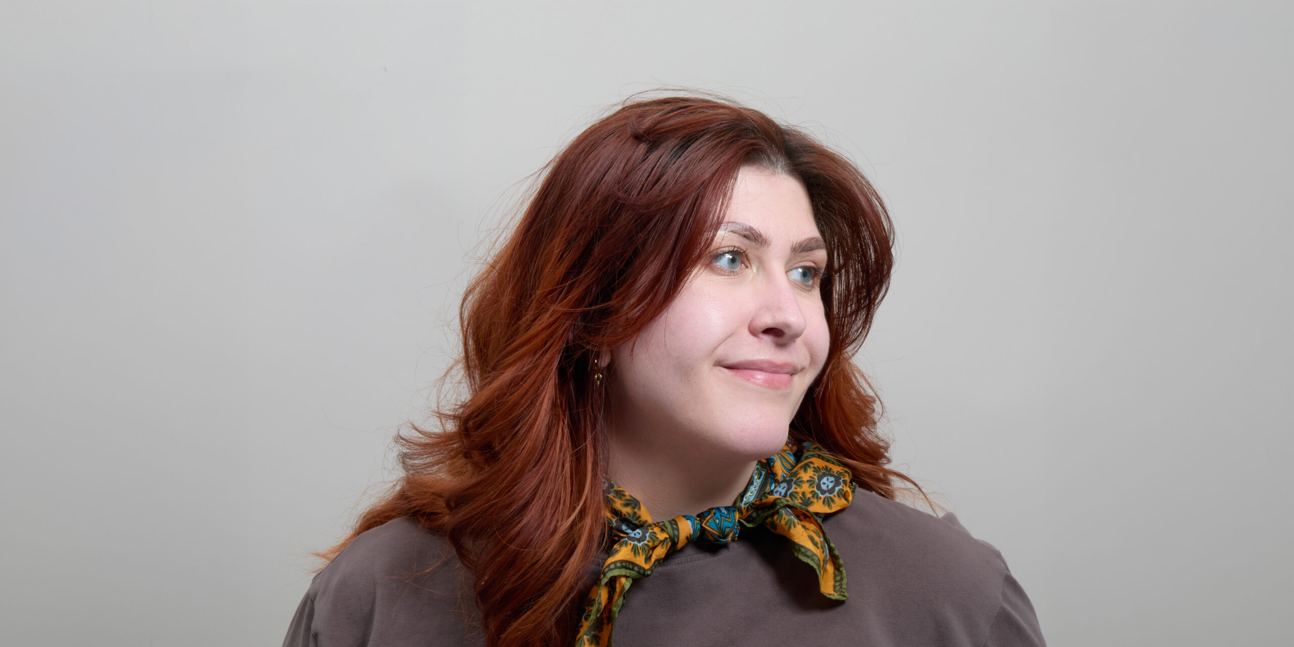 Cammie Niles with with long, wavy red hair smiles, wearing a patterned scarf and a gray top against a neutral background.