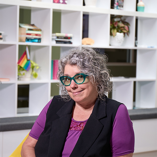 A woman with curly gray hair and glasses sits in front of a bookshelf, smiling slightly.