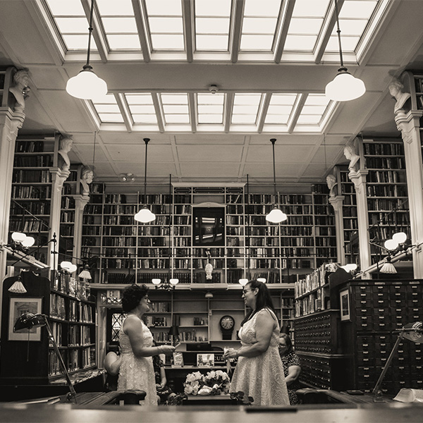 Two women in wedding dresses stand facing each other in a library filled with books.